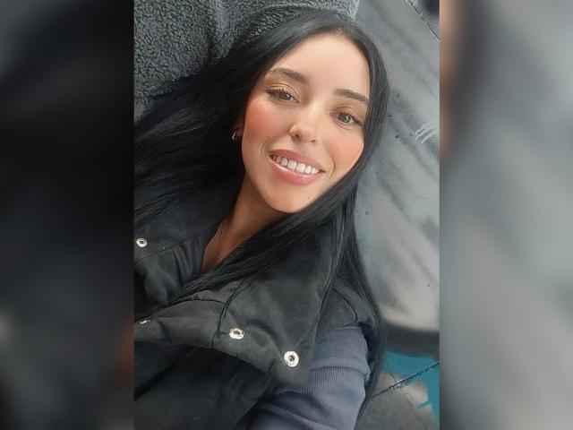 MartinaRey with long black hair, smiling, wearing a black jacket, has cute, small tits, and a cute nose, lying down, with a blurry gray background.