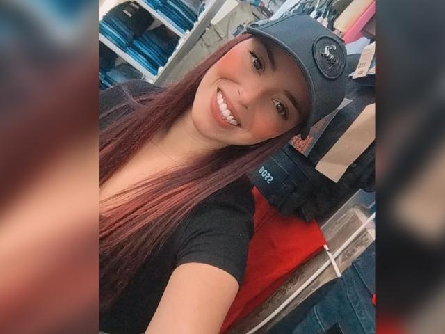 MartinaRey with long red-brown hair, smiling, wearing a black shirt and gray cap with a logo, standing in a store with blue and red items in the background.