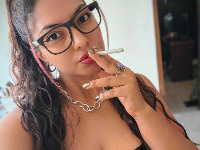 FetishWayraa with dark wavy hair, wearing black glasses, pink earbuds, chain necklace, and gold nail polish, smokes a cigarette, showing her shiny gold ring on her finger.