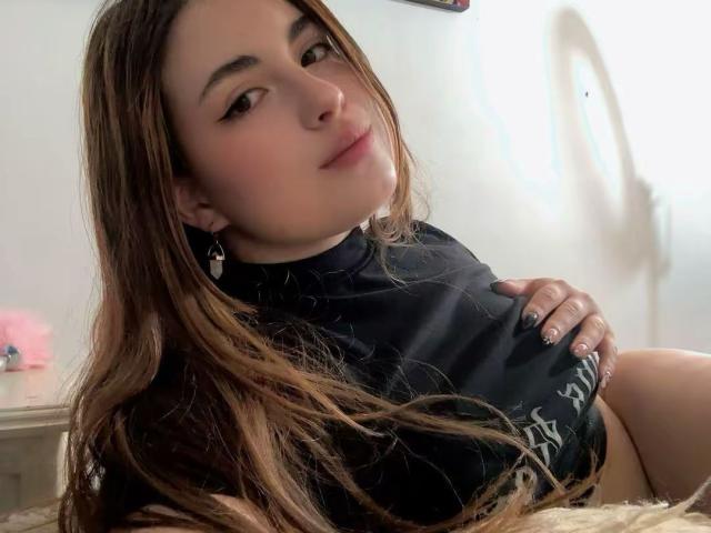VicluX with long brown hair, black top, holding her tits, slight smile, pink nail polish, white wall, ring light shadow, soft light, casual, relaxed, cute, confident.