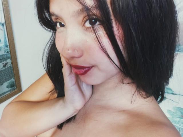 AbrilKoss with black hair, nose piercing, red lipstick, and hand on cheek, smiling, showing her tits, bare shoulders, soft light, cute, Asian, slight blush.
