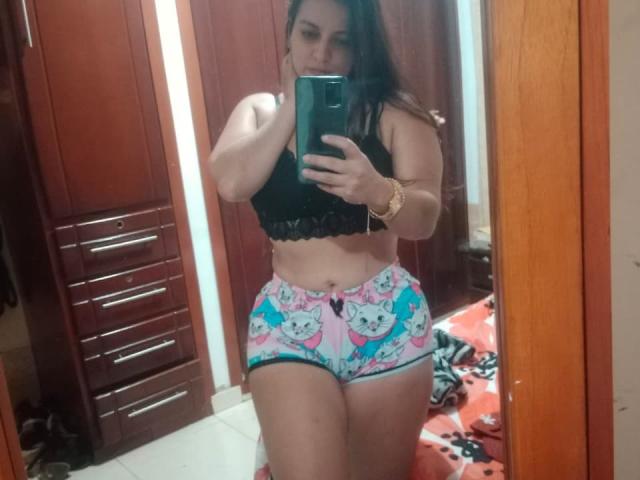 A selfie of LoveThickMonyque in a mirror. She has long dark hair, light brown skin, and a curvy physique. She wears a black lace bra and colorful, cartoon-printed shorts with a pink bow. Her right arm is raised, holding a black phone, and she wears a gold bracelet on her wrist. The background includes wooden closet doors and a red, patterned floor mat. The image is slightly blurred.