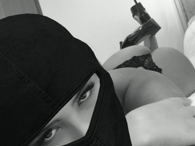 Black-and-white photo of AnnaPooky lying on her stomach, wearing a black mask covering her head, showing her eyes, and black lace panties, with her butt up in the air.