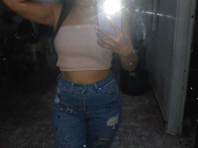 A selfie of SolaraDubois shows her from the chest down. She wears a pink ribbed, strapless top that exposes her midriff and a pair of blue, distressed jeans with multiple rips and patches. Her right arm is raised, holding a phone that flashes bright light, obscuring part of her face. The background is dark, with faint reflections on the mirror. The image is taken indoors, with a patterned floor visible. SolaraDubois has a slim, fit build.