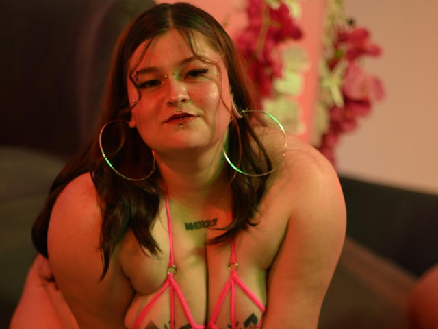 MilaDallila with pink strappy lingerie, large hoop earrings, nose ring, and lip piercing, leaning forward, showing her tits, with a tattoo above her cleavage, in red lighting.