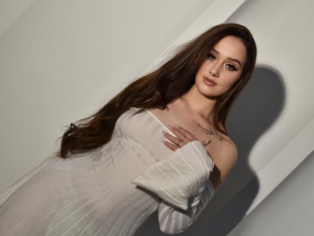 SabrinaCandi with long brown hair, wearing a sheer white dress, exposing small tits, hand on chest, tattoo above, soft shadow, light skin, looking at camera, subtle makeup.