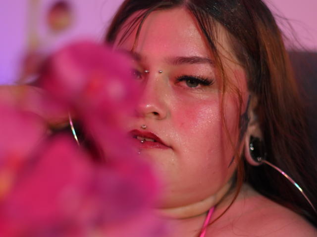 MilaDallila's face with a nose piercing, hoop earrings, and flushed skin, blurred pink flower in foreground, slightly parted red lips, and long brown hair, soft pink and purple lighting.