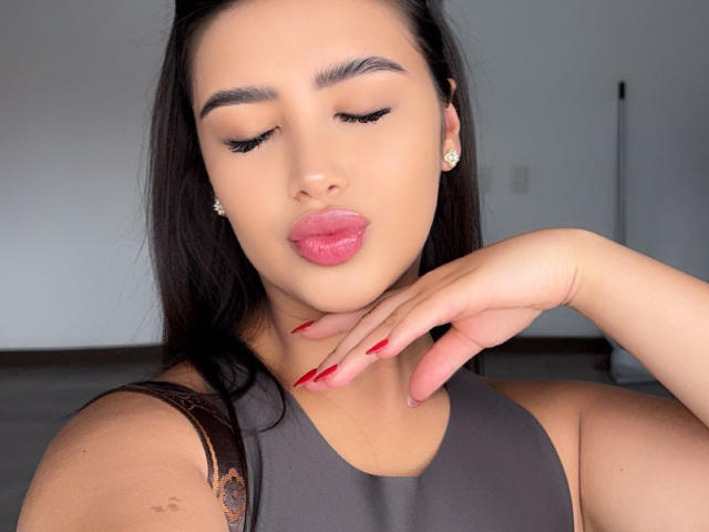 AnnaPooky with pouty pink lips, long red nails, dark brown hair, and cute earrings, wearing a gray sleeveless top, touching her chin.