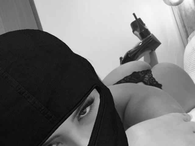 AnnaPooky, wearing a black mask, lies on her stomach, showing her ass in lace panties, with high heels and holding a gun, in a black-and-white selfie.