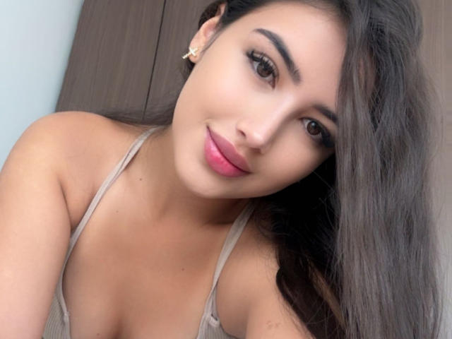 AnnaPooky has brown eyes, long dark hair, pink lipstick, and tan skin, wearing a beige top, showing her small tits and smiling at the camera.