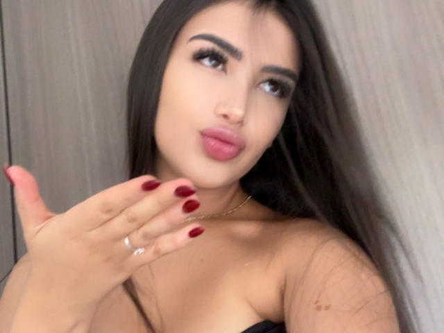 AnnaPooky, avec ses longs cheveux noirs, ses ongles rouges et son collier en or, regarde sur le côté avec ses lèvres roses pincées, et touche sa joue avec une bague à son doigt..