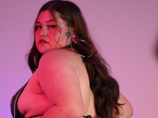 MilaDallila, a plus-sized woman with long, wavy brown hair, looks over her shoulder. She has a tattoo of a star on her left cheek, and large silver hoop earrings. Her skin is fair with red lighting highlighting her. She wears a black strap, revealing her back. Her expression is neutral, and she has a slight frown. The image has a purple-pink gradient background. Her right arm is slightly raised. The photograph emphasizes her upper body and facial features.