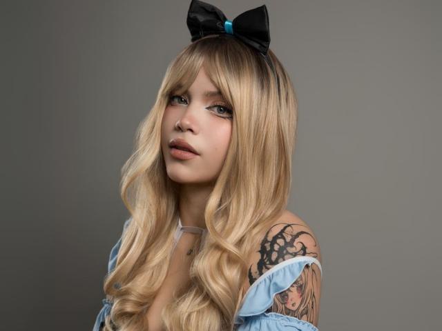 AryButty with blonde wavy hair, blue off-shoulder dress, black bow, and tattoos on her right shoulder, looking up with a slight smile. Her tits are partially visible.