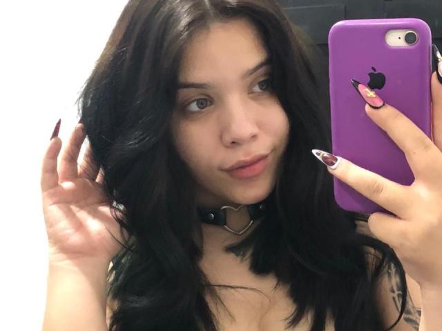 AryButty with dark wavy hair, wearing a black choker with a heart, holding a purple iPhone, and showing off her long, painted nails, looking at the mirror.