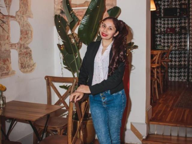 StephanieSun with curly dark brown hair, red lipstick, black blazer, white shirt, and blue jeans, leaning on wooden chair, next to large green plant, smiling.