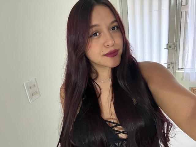 MarianaVargasX with long dark red hair, wearing a black lace-up top, showing her tits, purple lipstick, and a slight smile, taking a selfie in a bright room.