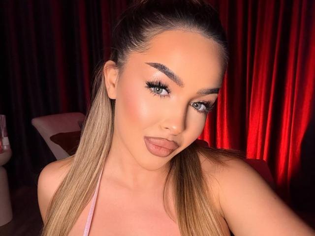 LisetteLuxe with long blonde hair, thick eyebrows, green eyes, and pink lipstick, sticking out her tongue. She's wearing a pink top, and her tits are slightly visible.
