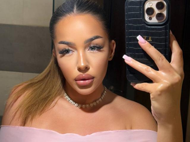 LisetteLuxe with shiny skin, full lips, and long nails holding a phone, wearing a pink off-shoulder top and pearl necklace, has her brown hair in a ponytail.