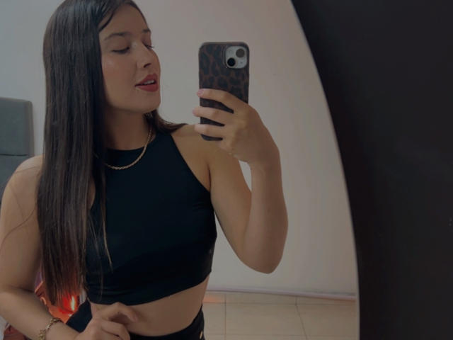EmmaLombardi takes a selfie in a mirror. She has long, straight brown hair, red lipstick, and a light skin tone. She's wearing a black sleeveless crop top that reveals her midriff and a black bottom. She holds a phone with a leopard print case in her right hand, slightly tilted. Her left hand rests on her hip. A gold necklace is visible. The mirror's reflection captures her from the waist up. The photo has a casual, modern style.