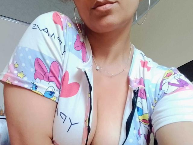 Close-up of Angelonix from the shoulders to the chest. Angelonix has light brown skin and wears a colorful, short-sleeved top with a deep V-neck, revealing significant cleavage. The top features pink bows, hearts, and stars on a white background with blue polka dots. Angelonix's lips are slightly parted, and she wears large silver hoop earrings and a delicate silver necklace. The image is brightly lit, highlighting the top's vibrant patterns and Angelonix's smooth skin.