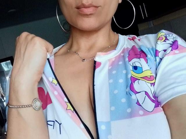 Angelonix with pink lips, hoop earrings, necklace, bracelet, and a colorful Disney-themed shirt, showing a deep V-neck, holding the shirt. Boobs partially visible.