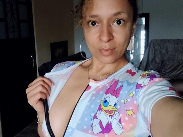Angelonix with curly brown hair, light brown skin, and small tits, wearing a colorful Disney-themed top, slightly open to reveal a black strap, holding it with a finger.