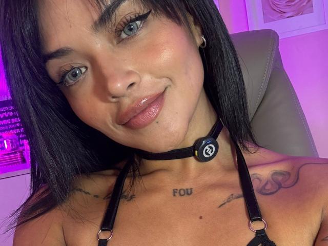 MajoJones with black hair, blue eyes, and a cute smile, wearing a black choker with a logo, and showing tattoos on her chest and shoulders, with shiny pink lips.