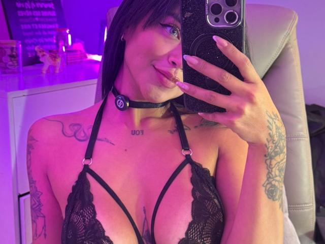 MajoJones taking a selfie, wearing black lace bra, choker, tattoos, purple lighting, sticking tongue out, holding phone, visible large tits, long nails, chair in background.