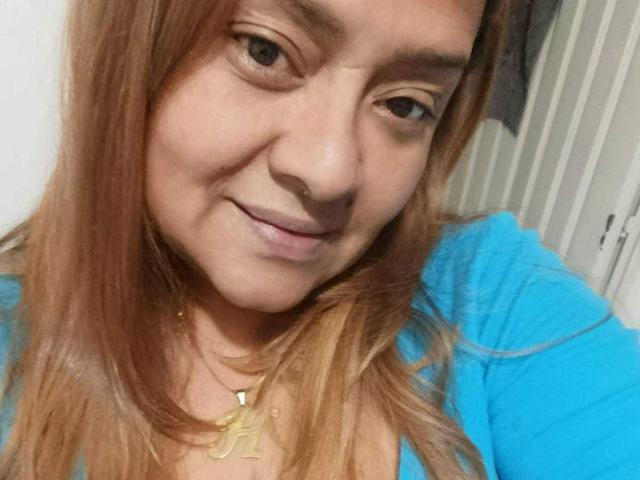 MonzerratBoyer with light brown hair, blue shirt, gold necklace, and subtle smile, showing her face and upper chest in a selfie, looking slightly to the side.