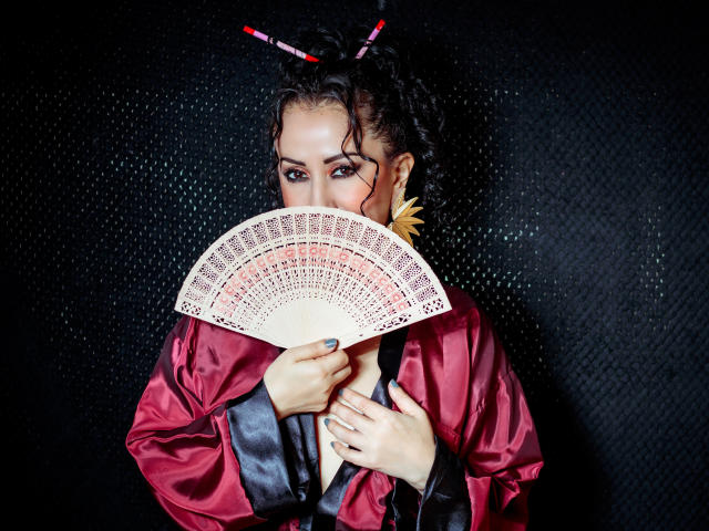 SamanthaAntolin holds a fan with intricate red and white patterns in front of her face. She has dark, curly hair styled with red and pink hair sticks and wears a shiny red satin robe with black trim. Her eyes are focused, and she has dark, dramatic makeup. A large, yellow feather earring is visible on her right ear. The image is sharp, with a dark, textured background.
