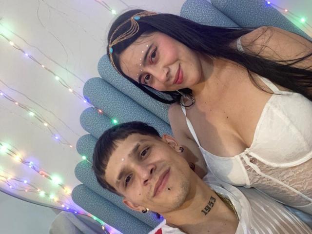 A selfie of AnjaliRohan and a man. AnjaliRohan has long black hair, fair skin, and a headband with gold details. She wears a white lace-up corset that reveals her cleavage. The man has short dark hair, a small tattoo under his chin, and black ear gauges. They both have small, white facial markings. The man is in a white shirt. Multicolored string lights are in the background. AnjaliRohan is on the right, the man is on the left.