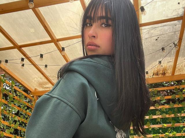 MiaRousel with long black hair, green hoodie, and pouty lips, looking over shoulder, under wooden pergola with string lights, has a sexy, cute, and attractive face.