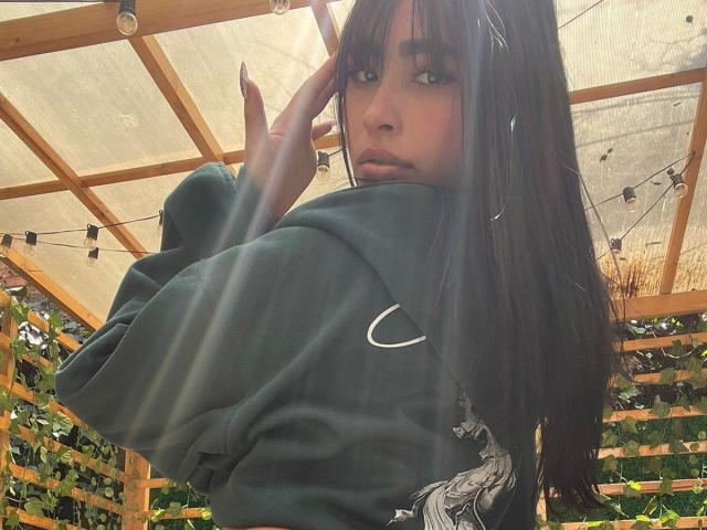 MiaRousel with long black hair, green Nike sweatshirt, looking over shoulder, hand in hair, wooden pergola, string lights, greenery, soft light, serious expression, dark eyes, slight pout.