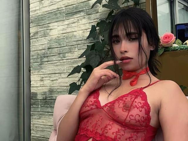 MiaRousel with dark hair and bangs, wearing red lace lingerie, red choker, and pink rose, biting her finger, sitting in a chair, looking at the camera.
