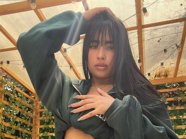 MiaRousel with long black hair, green hoodie, hand on chest, exposing her tits, under wooden canopy with string lights, looking at camera, pink nails, serious expression.