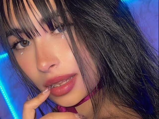MiaRousel's face with dark, straight hair, blue eyes, and pink lips, biting her finger, wearing a pink choker, and small beauty marks near her eyes.