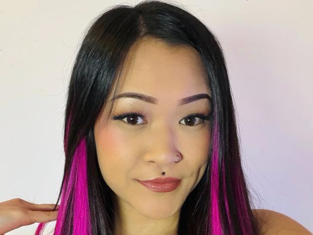 AsunaBloom-hot has long black hair with pink highlights, a nose ring, and a lip piercing. She's smiling with bold eyeliner and pink lipstick.