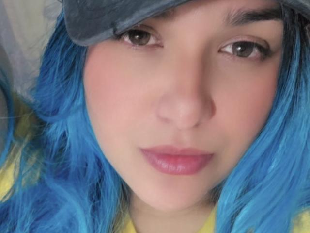 Close-up of VivianaThompson with blue hair, brown eyes, and pink lips. She wears a gray cap and a yellow shirt. Her skin is light, and her eyebrows are dark. The blue hair contrasts with the yellow shirt and gray cap. VivianaThompson's face is centered, with a slight smile. The image is sharp, focusing on her facial features. Her eyes are slightly up, and her lips are slightly parted.