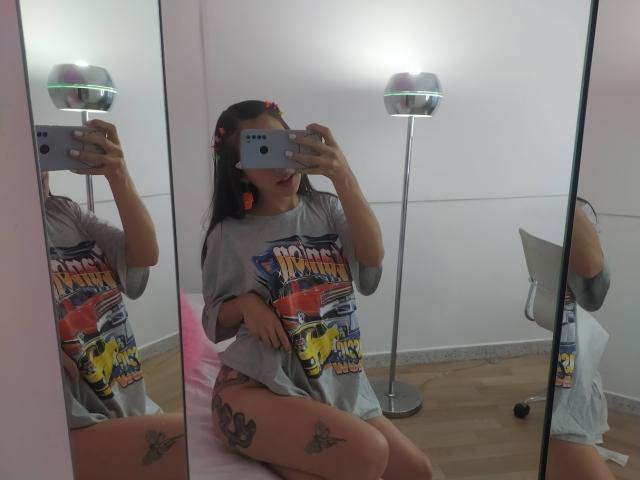 CattalinaBotero in a grey "Van Halen" shirt, sitting on a pink bed, taking a mirror selfie, showing tattoos on thighs, with a phone and a lamp in the room.