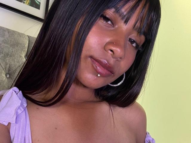 NahomyNoir has dark brown skin, long straight black hair with bangs, a nose ring, a lip piercing, and wears a purple off-shoulder top with a hoop earring.