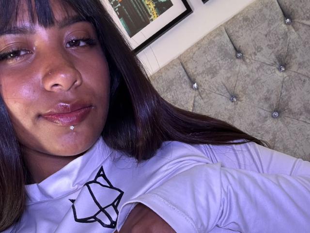 NahomyNoir with dark brown hair, nose ring, and lower lip piercing, wearing a white shirt with a black geometric design, smiling slightly, lying on a tufted grey headboard.