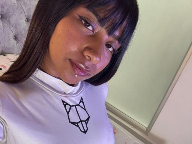 NahomyNoir has dark brown hair, glossy pink lips, and a white turtleneck with a black fox head graphic. Her face has a shiny, wet look.