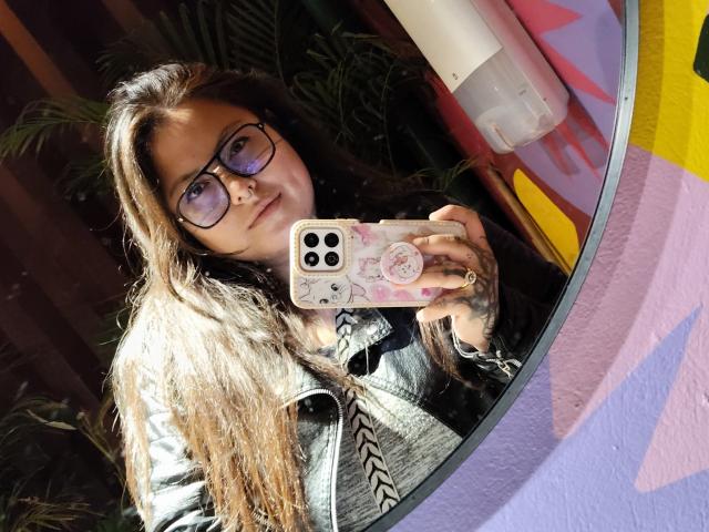 LilithHeavy with long brown hair, glasses, and a grey jacket, holding a pink phone with a sticker, taking a mirror selfie, showing tattoos on her fingers.
