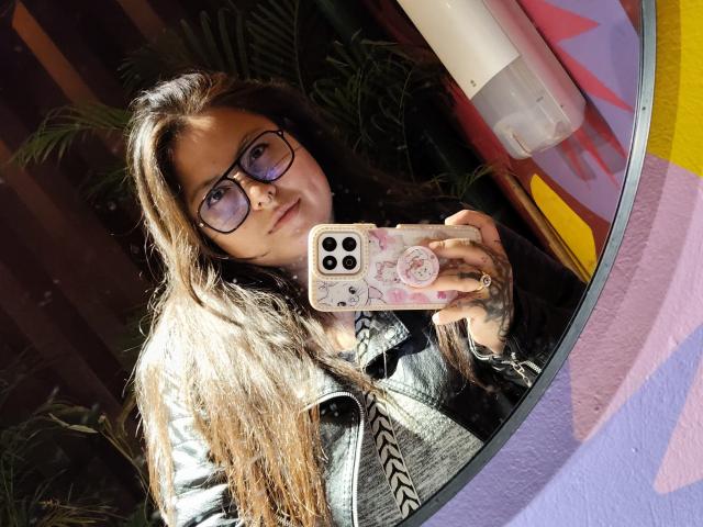 LilithHeavy with long brown hair, glasses, and a silver jacket, holding a pink phone with a pop socket, taking a mirror selfie, showing off her tits.