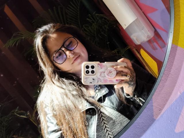 LilithHeavy with long brown hair, glasses, and a denim jacket, taking a mirror selfie with a pink phone case, showing tattoos on her hand.