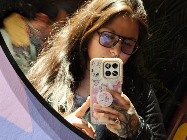 Lilith, con el pelo castaño ondulado, gafas y tatuajes, está tomando un selfie con un teléfono cubierto de pegatinas de gatos. Lleva una chaqueta de cuero negra y está mirando hacia abajo..