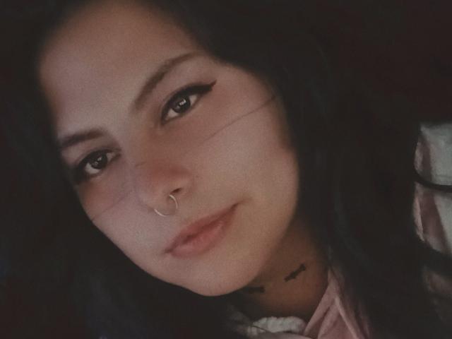 LilithHeavy has dark wavy hair, a nose ring, and black eyeliner with a small smile. Her skin is light, and she has a tattoo under her chin.