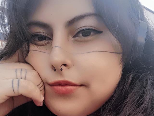 LilithHeavy has black eyeliner, a septum piercing, and black tattoos on her face and hand. She's resting her head on her hand with red lipstick.