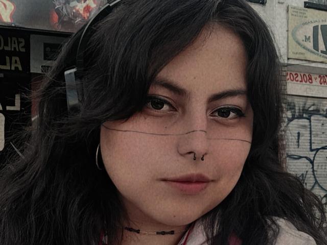 LilithHeavy has dark wavy hair, brown eyes, a nose ring, and a thin line under each eye. She's wearing headphones and a choker, with a slight, confident smile.