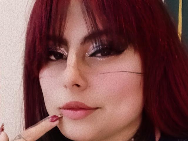 LilithHeavy with red hair, black eyeliner, and pink lipstick, biting her finger with red nails, showing a small scar on her right cheek. Her expression is slightly smug.
